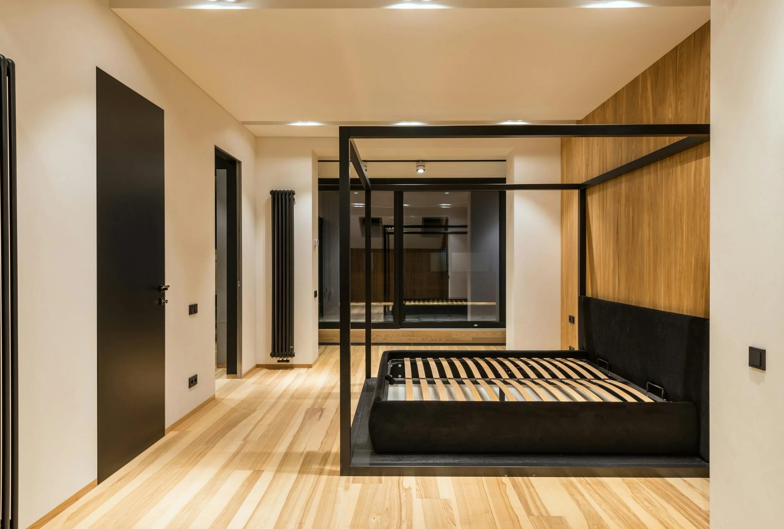 Modern bedroom with a black bed frame and mattress, wooden floor, beige walls, black door, black radiator, and large window with black framing and a radiator below.