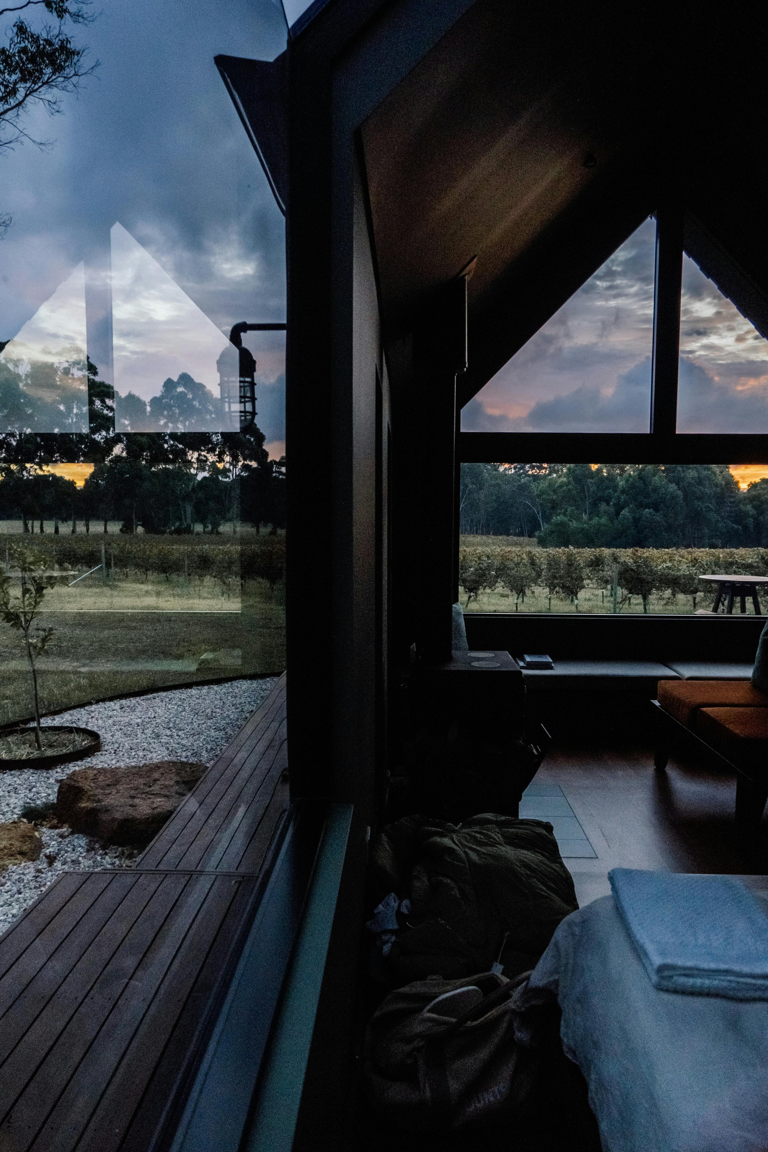 Inside a room with large glass windows showing an outdoor landscape of trees and a vineyard at sunset, with a couch, a table, and some bags on the floor.
