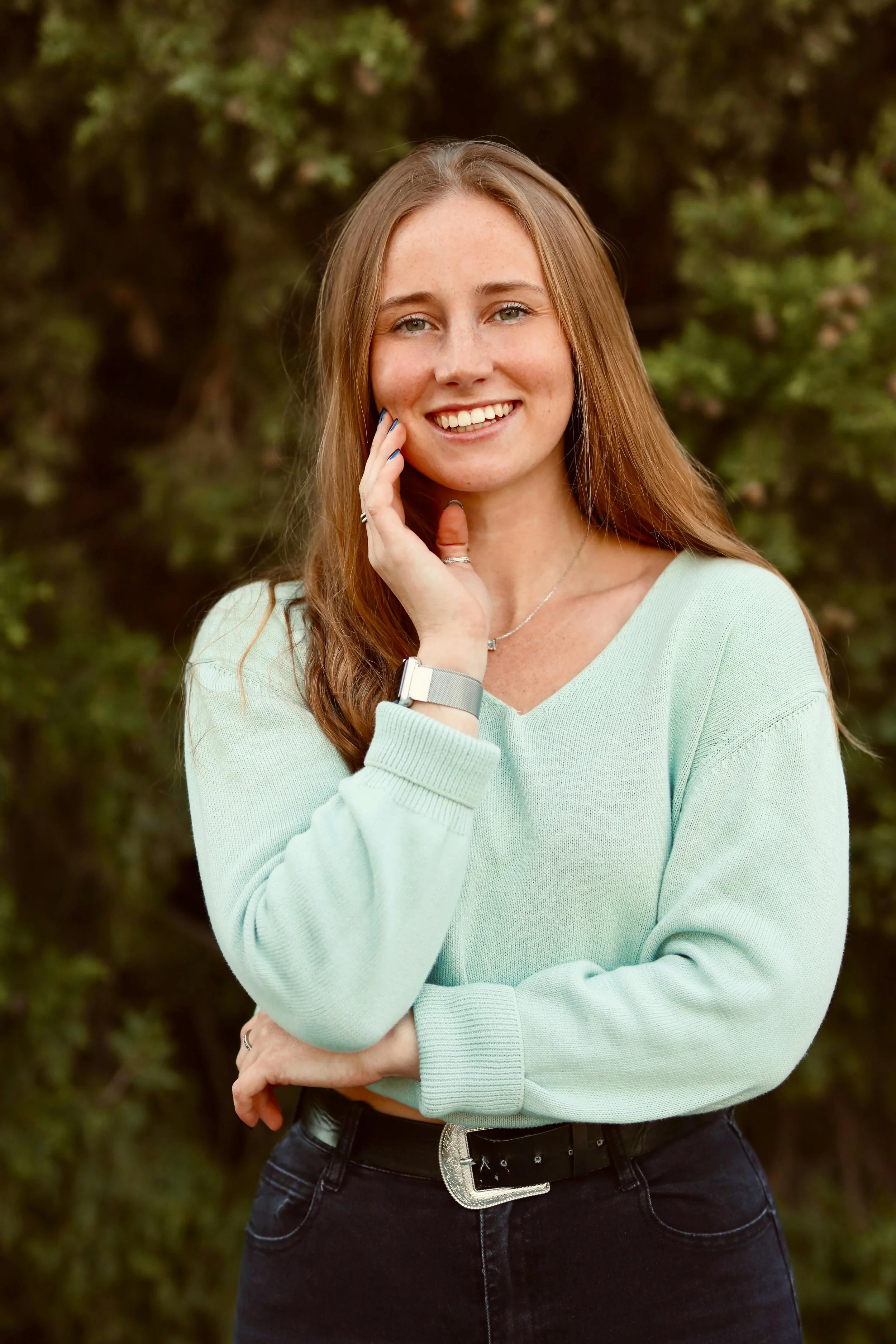 A young woman with long reddish-brown hair, smiling, standing outdoors in front of green shrubbery, wearing a light mint green sweater, black jeans, and a watch.