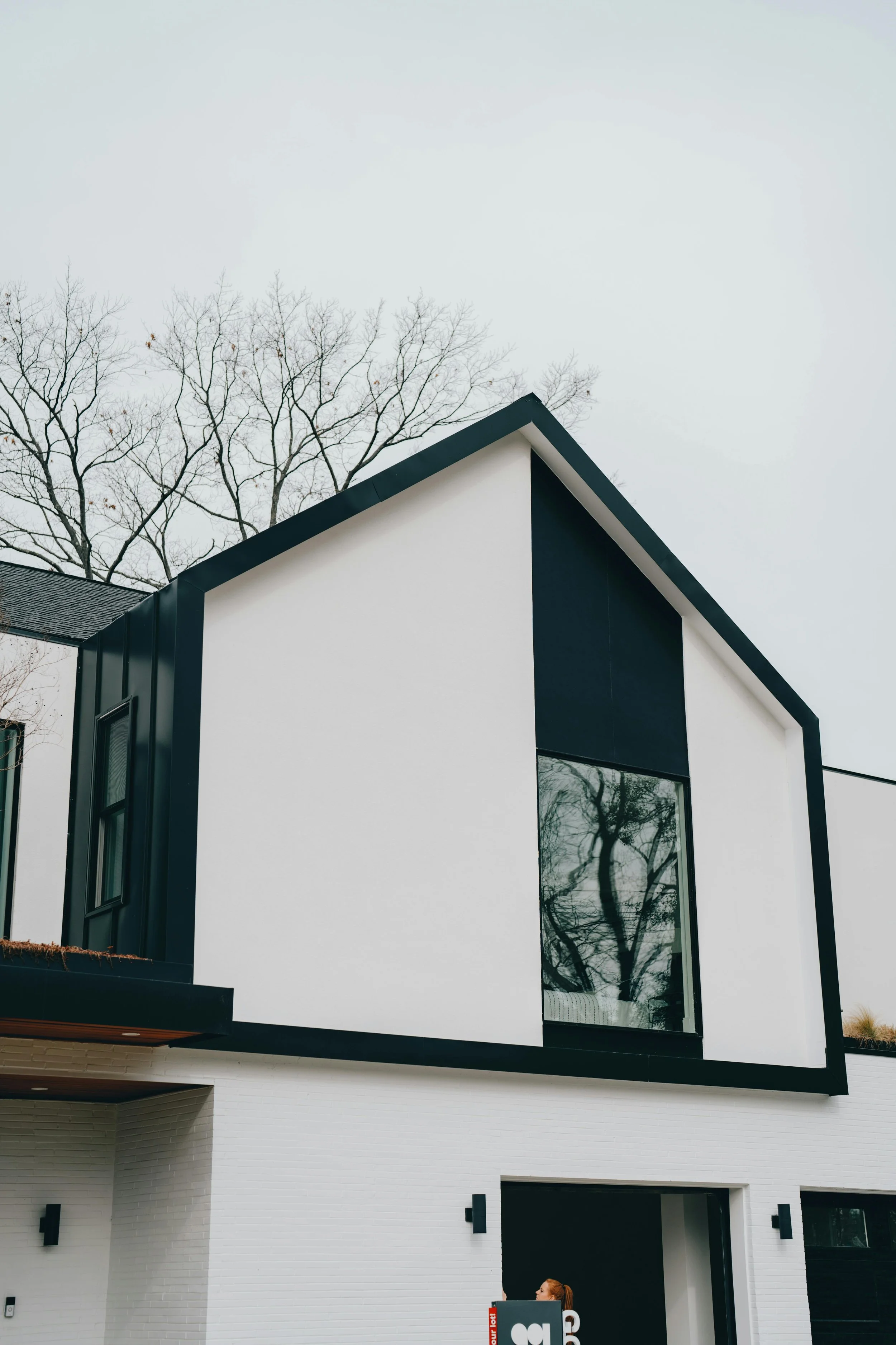 Modern white and black house with large windows and a woman holding a sign near the entrance, background of leafless trees and a cloudy sky.