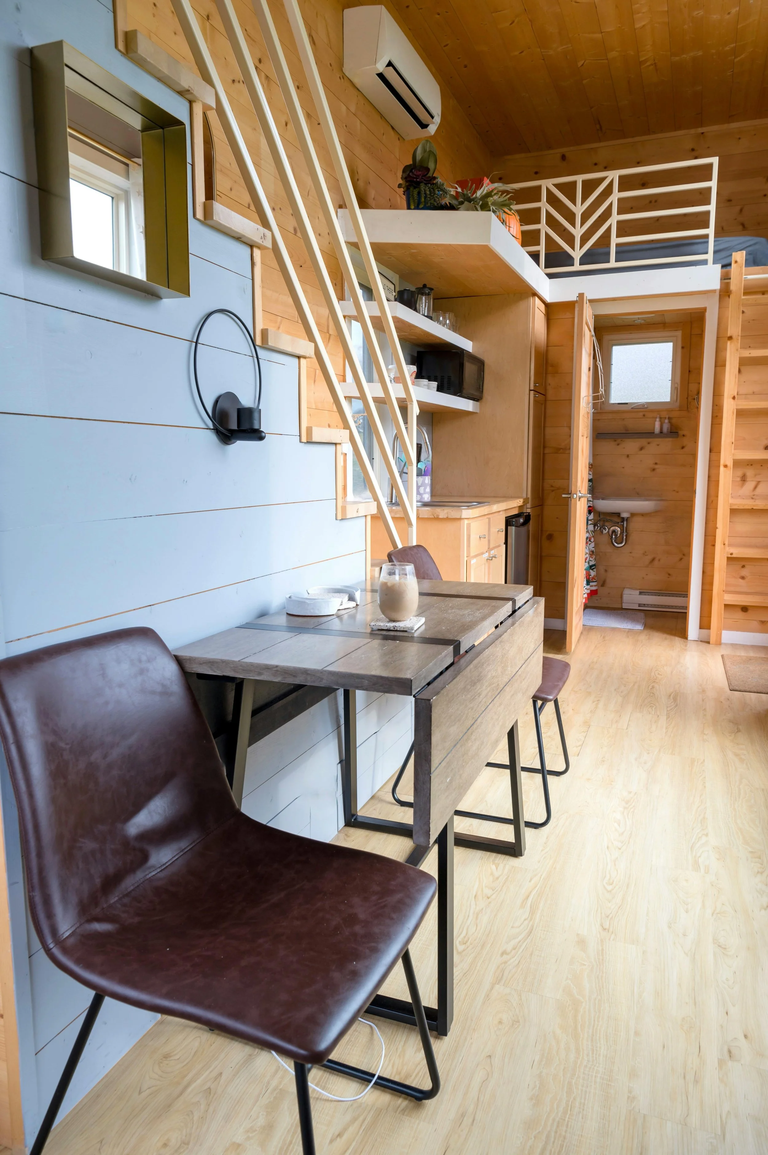 Interior of a small wooden cabin with a breakfast table, chairs, a mini kitchen area, and a loft space. The walls are wooden, and there is a window with an air conditioning unit above.