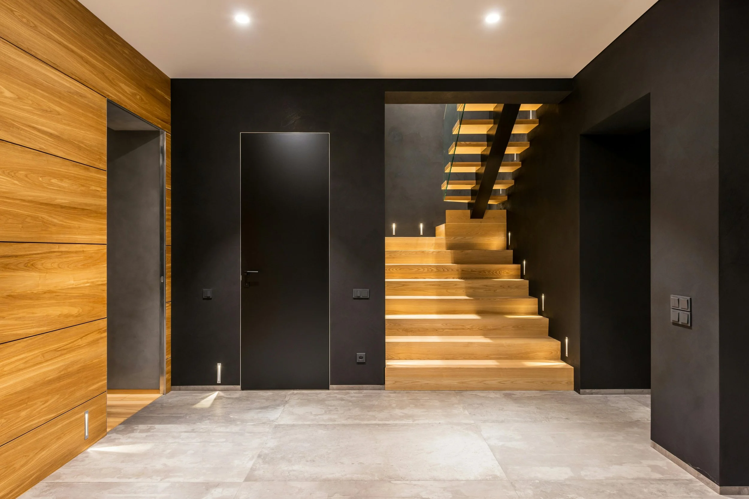 Modern interior with black and wood walls, a closed black door, grey tiled floor, and a wooden staircase with lighting.