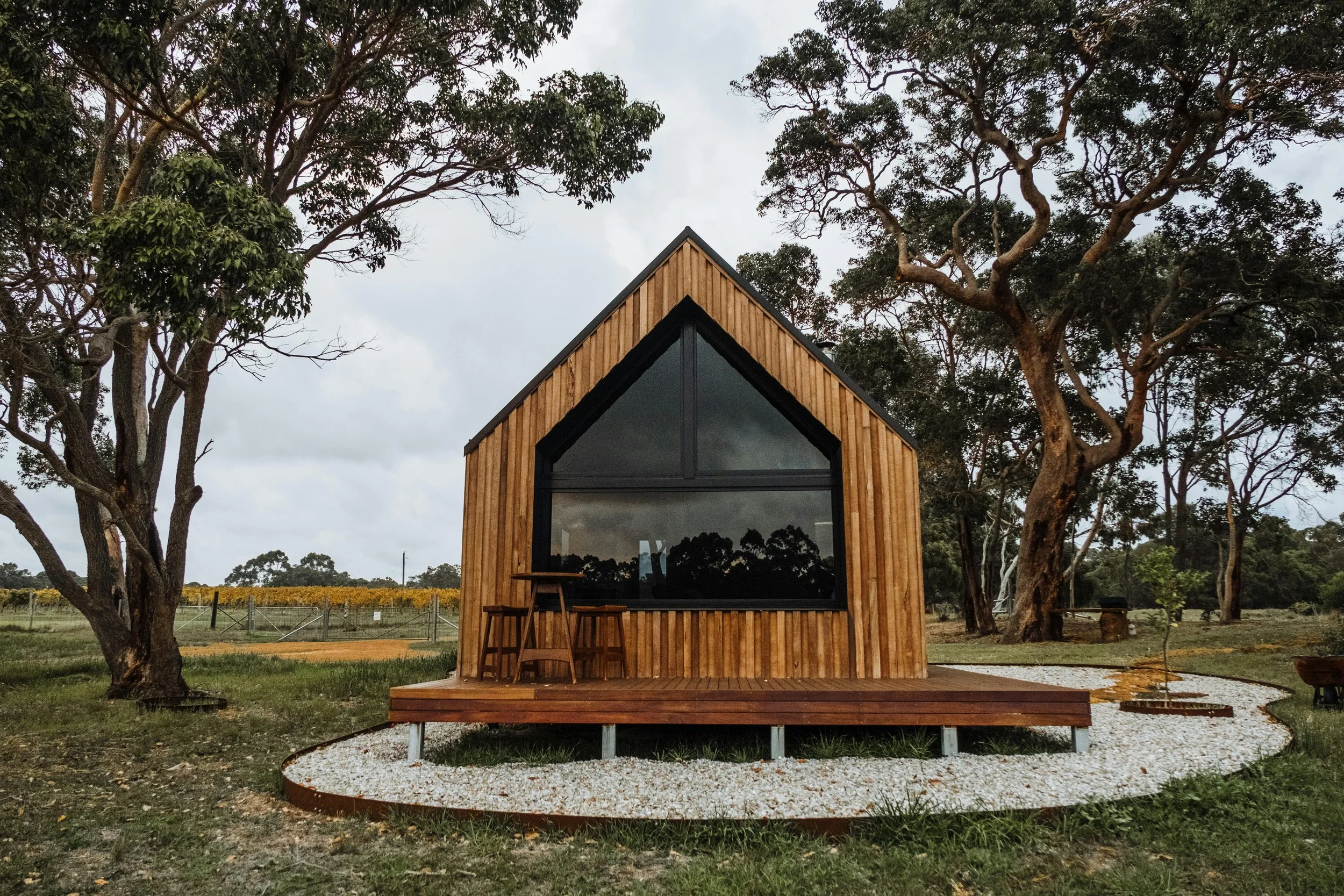 A modern tiny house with a triangular roof and large front window, built with wooden panels, situated outdoors near trees and a gravel pathway.