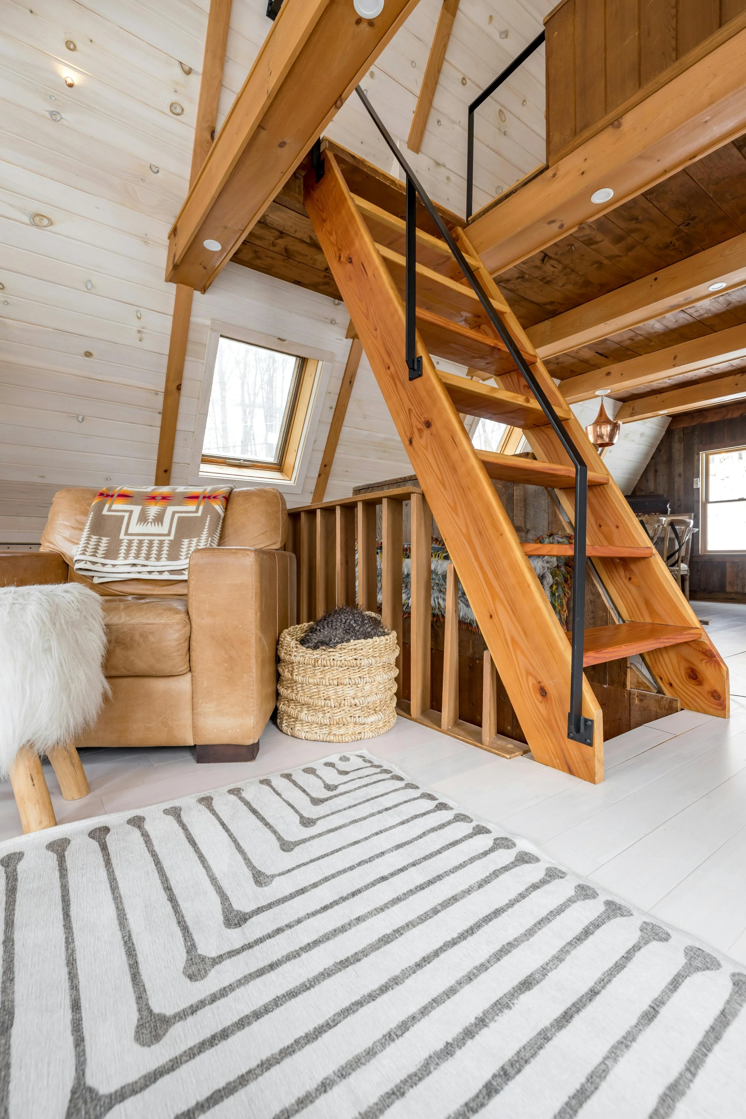 Interior of a cozy attic room featuring a wooden staircase, a leather armchair with a southwestern-patterned blanket, a woven basket with a blanket, a patterned rug, and skylight windows in a rustic style.