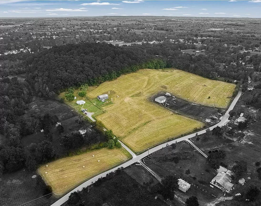An aerial view of a rural area with a large, open grassy field, scattered trees, and a few buildings, surrounded by a mix of forest and residential neighborhoods.