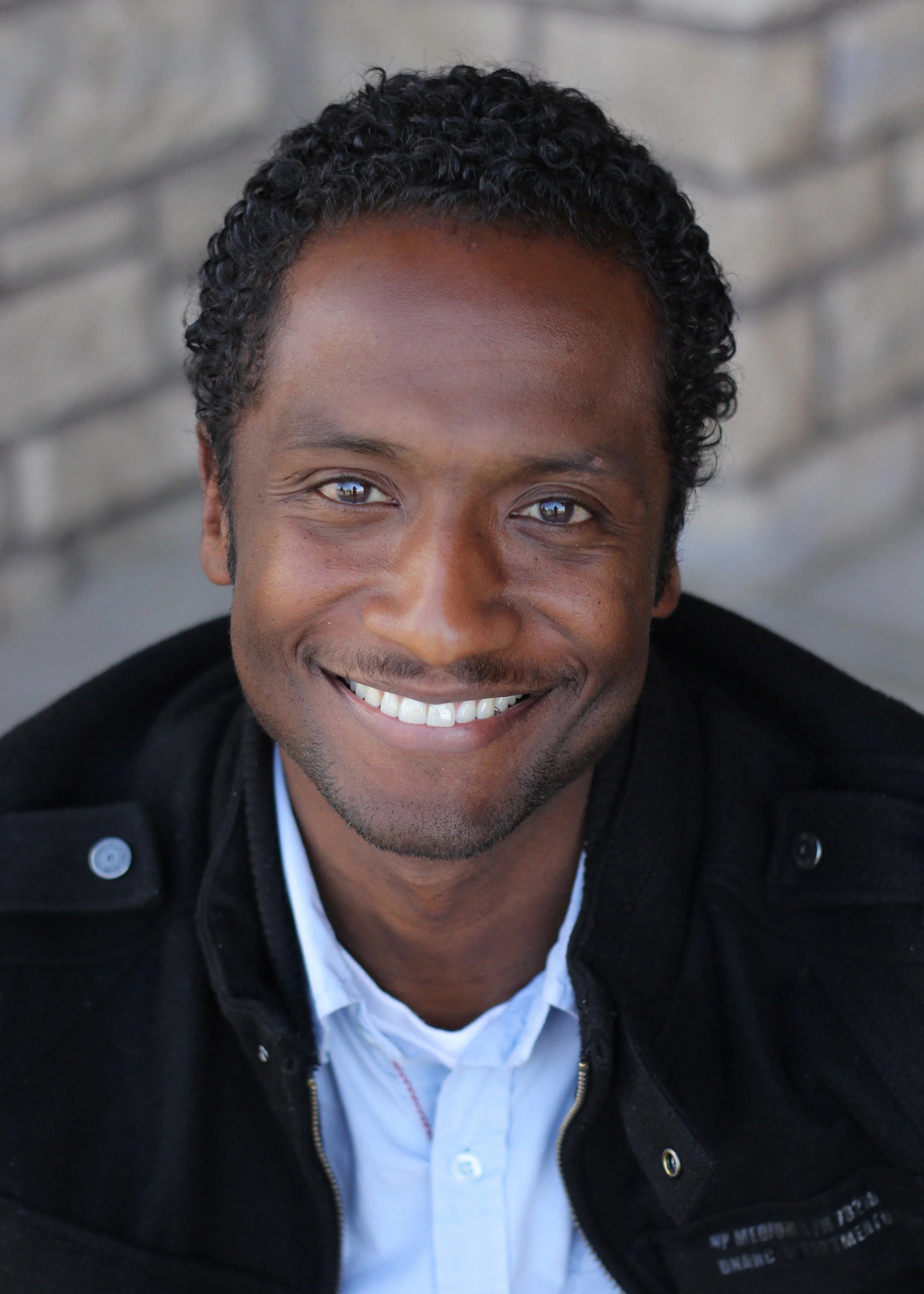 Portrait of a smiling man with curly black hair, wearing a black jacket and white collared shirt, in front of a blurred brick wall background.