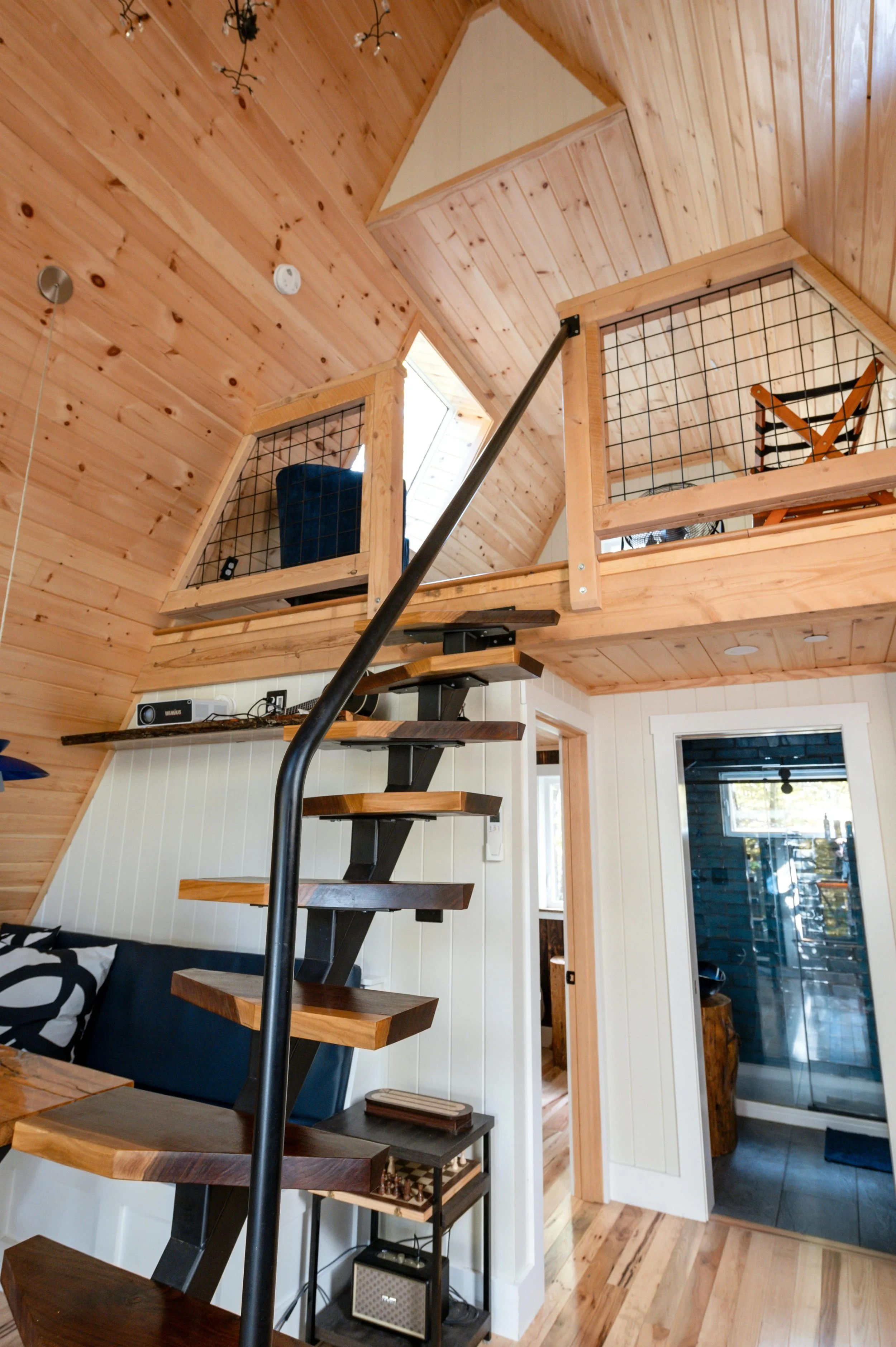Interior of a cozy wooden cabin with a staircase leading to a loft area, featuring wood paneling on the walls and ceiling, a small door to a bathroom or closet, and a sliding glass door leading outside.