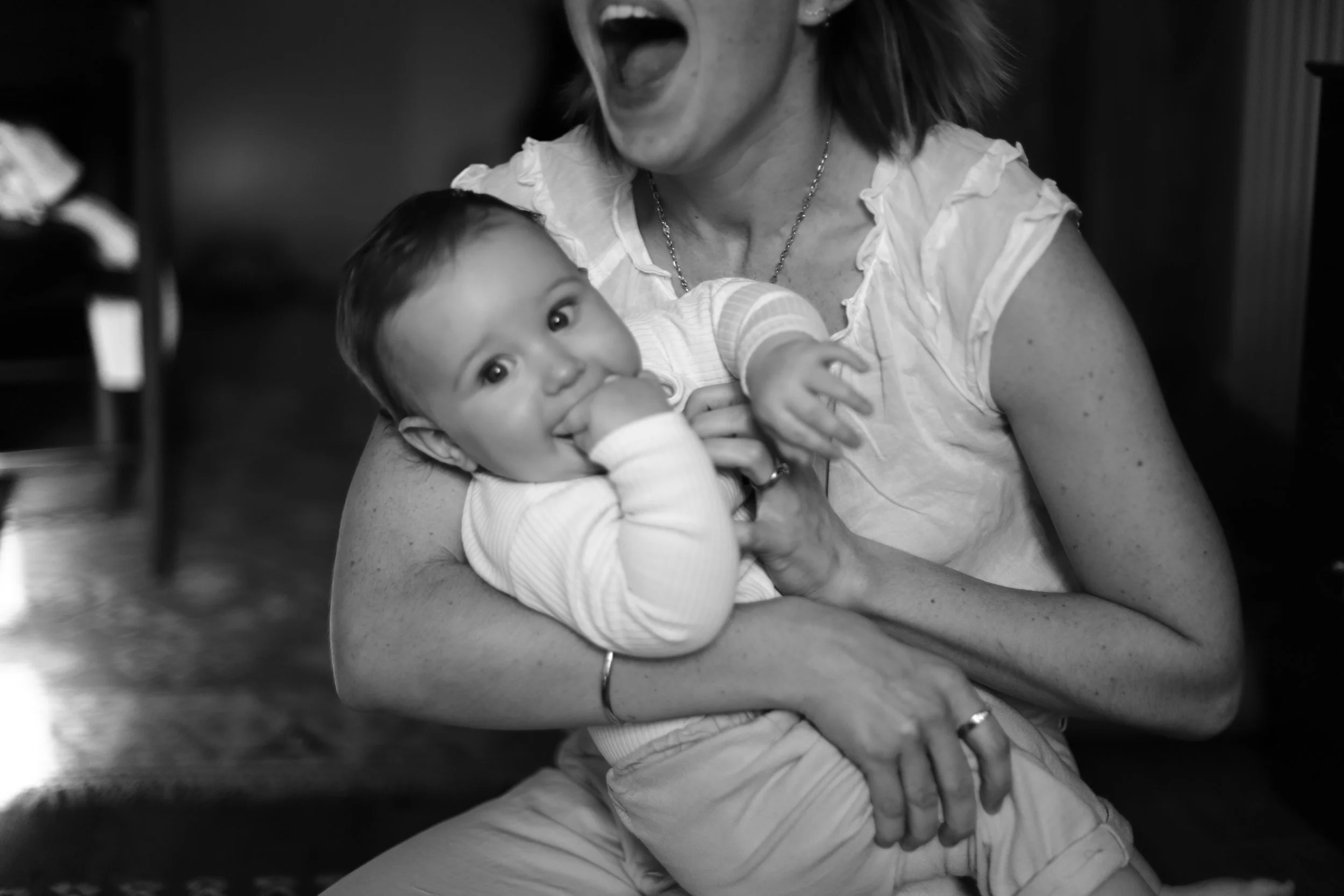 A woman is holding a smiling baby in her arms, the baby has one hand in her mouth and the woman is smiling.