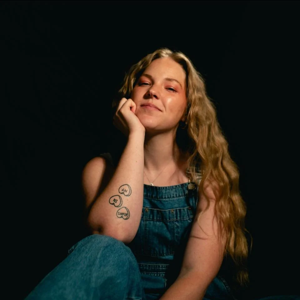 A young woman with long wavy ginger hair, wearing denim overalls, is seated against a black background. She has a tattoo on her right forearm consisting of five speech bubbles with the words 'Hello,' 'Bye,' 'Thanks,' 'Sorry,' and 'Sorry' inside. She is resting her chin on her right hand, with a slight smile and a playful expression.