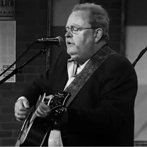 A man with glasses and light-colored hair singing and playing an acoustic guitar with a strap, in a black blazer, in a performance setting.