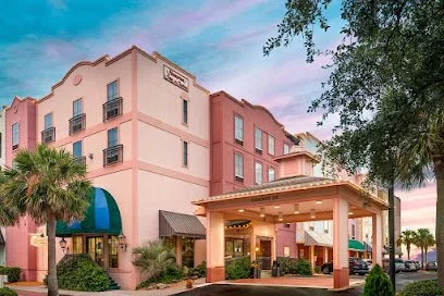 A pink hotel with a covered entrance, blue awning, palm trees, and outdoor seating, under a colorful sky.