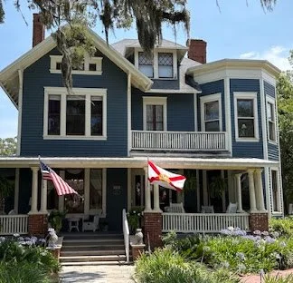 Large blue Victorian-style house with white trim, a wraparound porch, and American flags. Surrounded by greenery and trees.