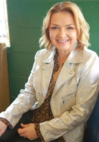 Smiling woman with blonde hair wearing a beige leather jacket sitting indoors with a green wall in the background.