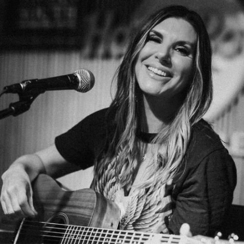 A woman smiling and playing an acoustic guitar on stage with a microphone.
