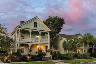 A large, two-story house with a wraparound porch, illuminated windows, surrounded by palm trees and bushes, under a sky with pink and purple clouds at dusk.