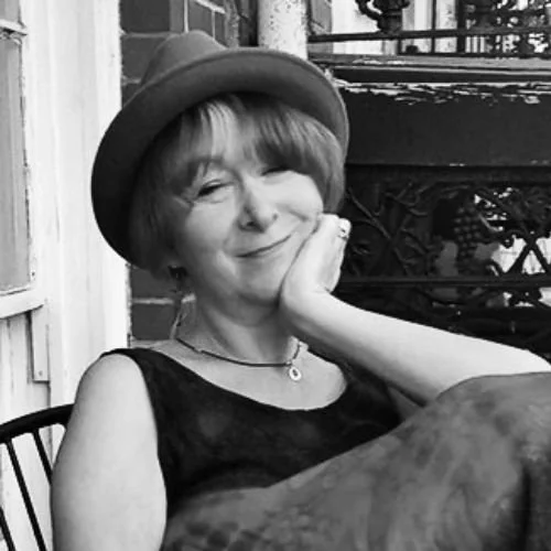 A woman with short hair wearing a hat, sitting outdoors, resting her cheek on her hand, and smiling at the camera.