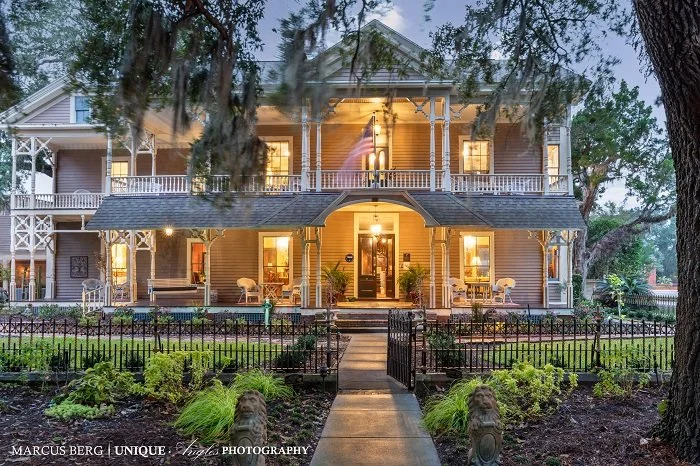 Large Victorian-style house with a front porch, lit windows, and a well-maintained garden, taken during dusk.