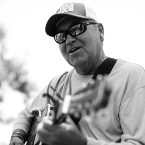 A man wearing sunglasses, a cap, and a light-colored shirt, playing an acoustic guitar outdoors.