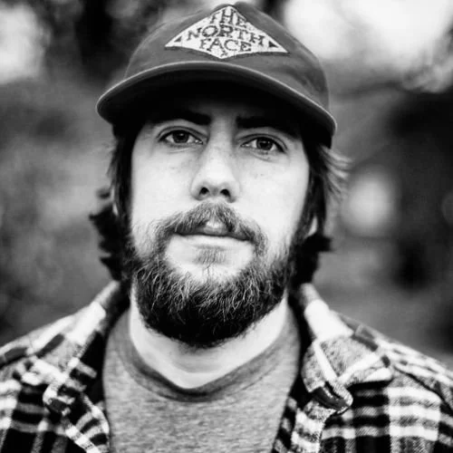 Portrait of a man with a beard, wearing a baseball cap and a plaid shirt, outdoors in black and white.
