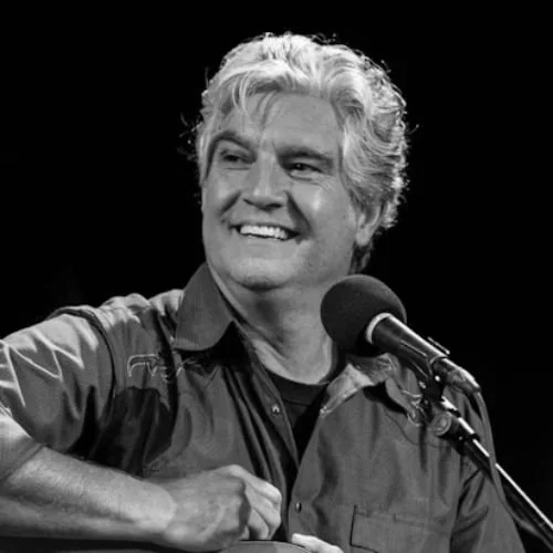 Black and white photo of a man smiling and playing guitar with a microphone in front of him.