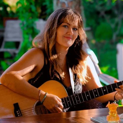 Woman sitting outdoors playing an acoustic guitar, smiling at the camera, greenery in the background.