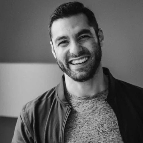 A smiling man with dark hair and a beard, wearing a jacket over a t-shirt, standing indoors.