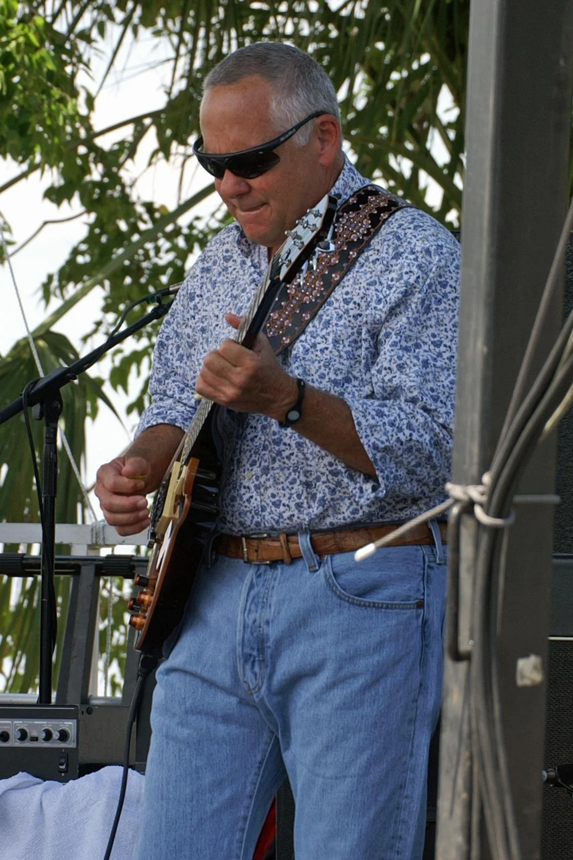 A man wearing sunglasses and a floral shirt playing an acoustic guitar outdoors among green trees.