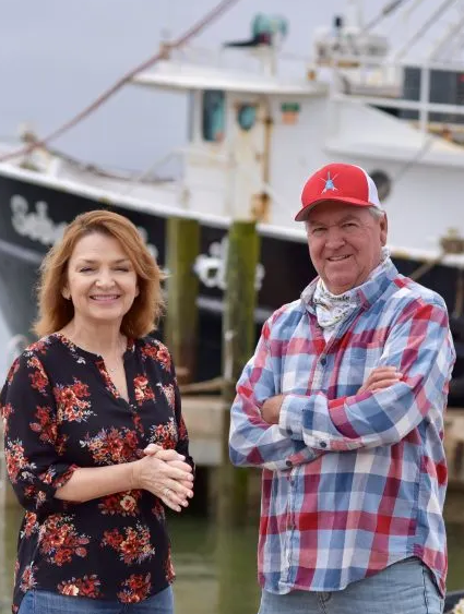 A woman and a man standing near boats at a marina. The woman is smiling with her hands clasped, wearing a floral blouse. The man is smiling with arms crossed, wearing a plaid shirt, a bandana around his neck, and a red cap. In the background, there are boats docked at the marina.