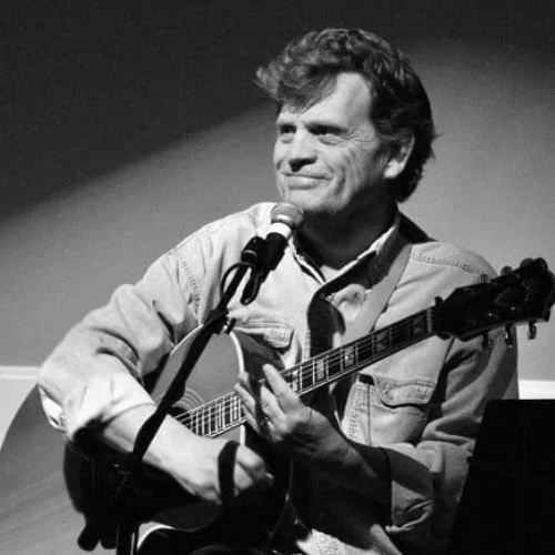 A man with wavy hair playing an acoustic guitar and singing into a microphone on stage.