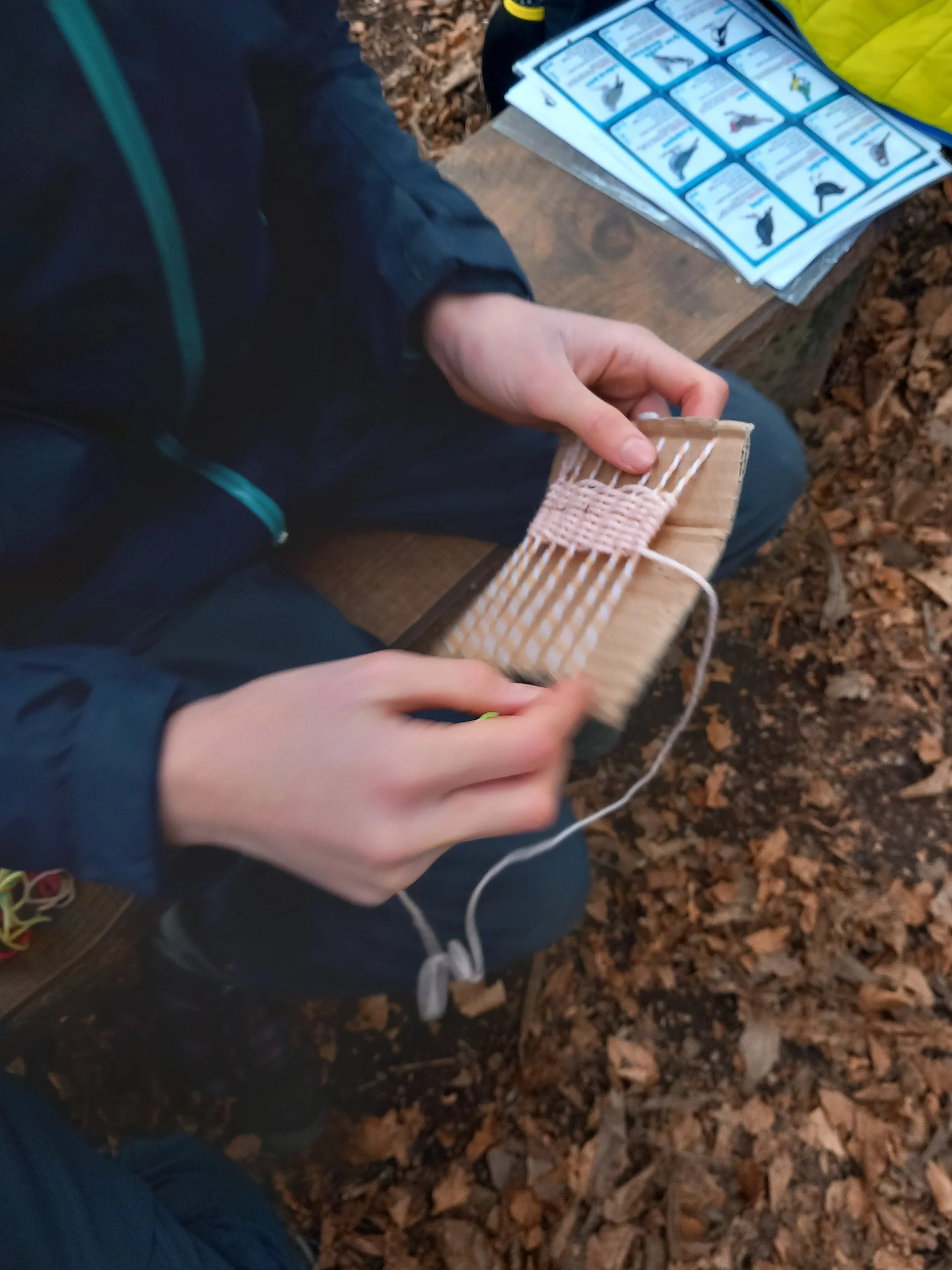 A person sitting on a wooden bench in a forest, crafting a woven bracelet using a small cardboard piece, pink thread, and a crochet hook; an instruction sheet with a series of bird illustrations lies on the bench.