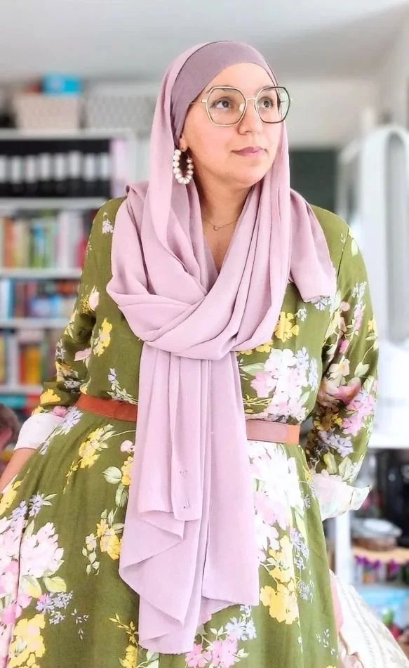 A woman wearing glasses, a purple headscarf, and a floral dress with an olive green background in an indoor setting with bookshelves in the background.