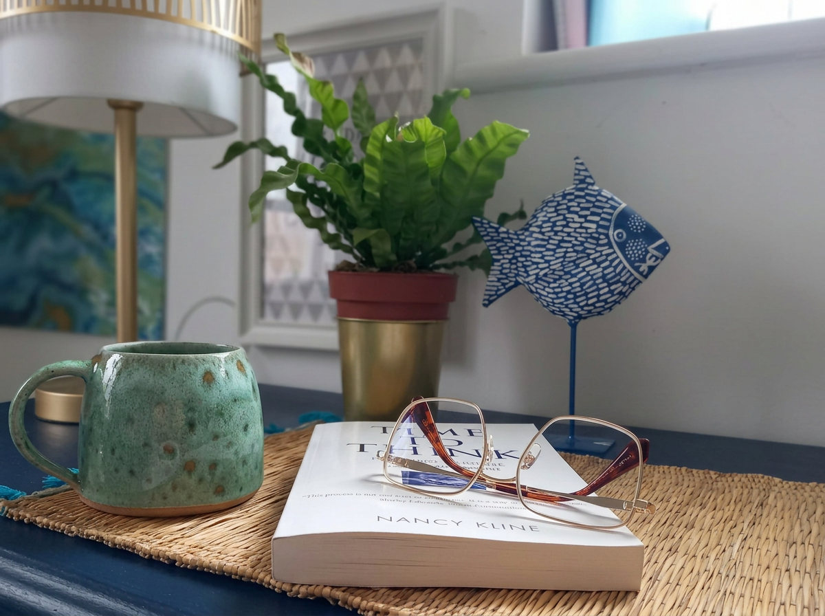 A cozy tabletop setup with a green ceramic mug, a pair of sunglasses, a book titled 'Time to Think' by Nancy Kline, a potted plant with lush green leaves, and a decorative blue fish sculpture on a woven mat near a window.