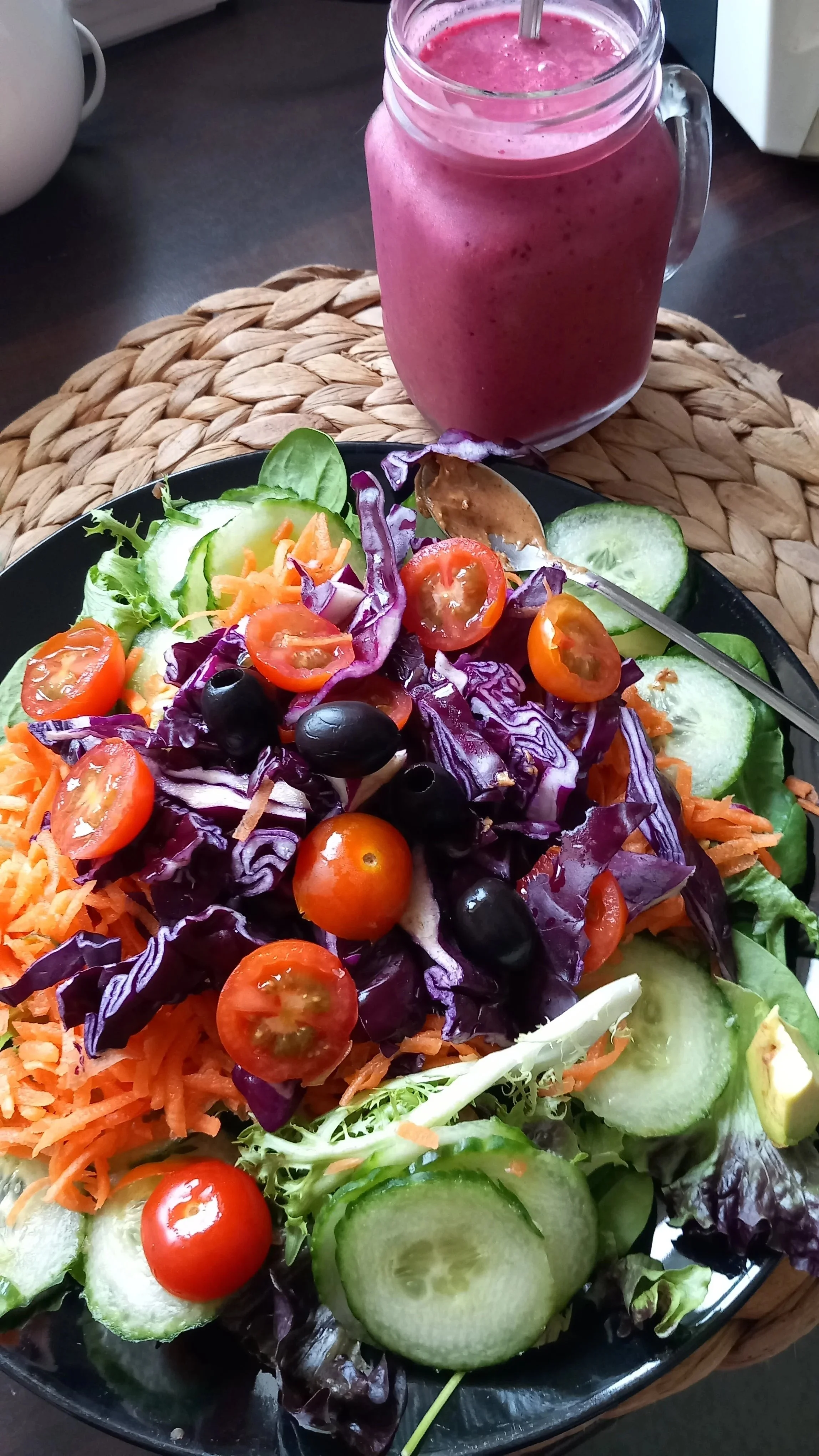 A colorful salad with cherry tomatoes, cucumber slices, purple cabbage, shredded carrots, black olives, and green lettuce on a black plate. In the background, a pink smoothie in a mason jar with a metal straw.