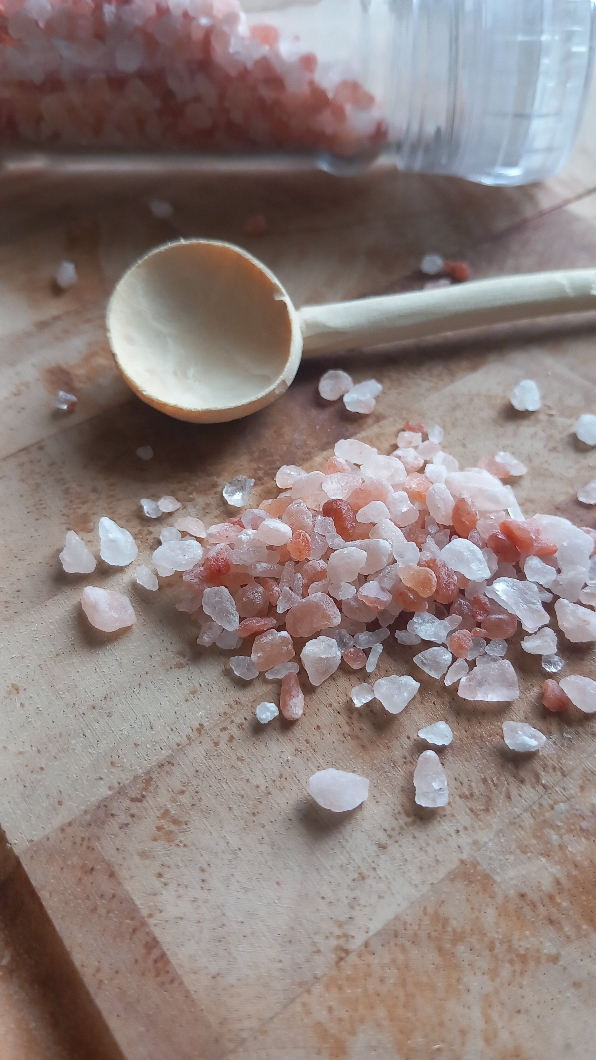 Pink Himalayan salt crystals scattered on a wooden surface with a spoon and a container of salt in the background.