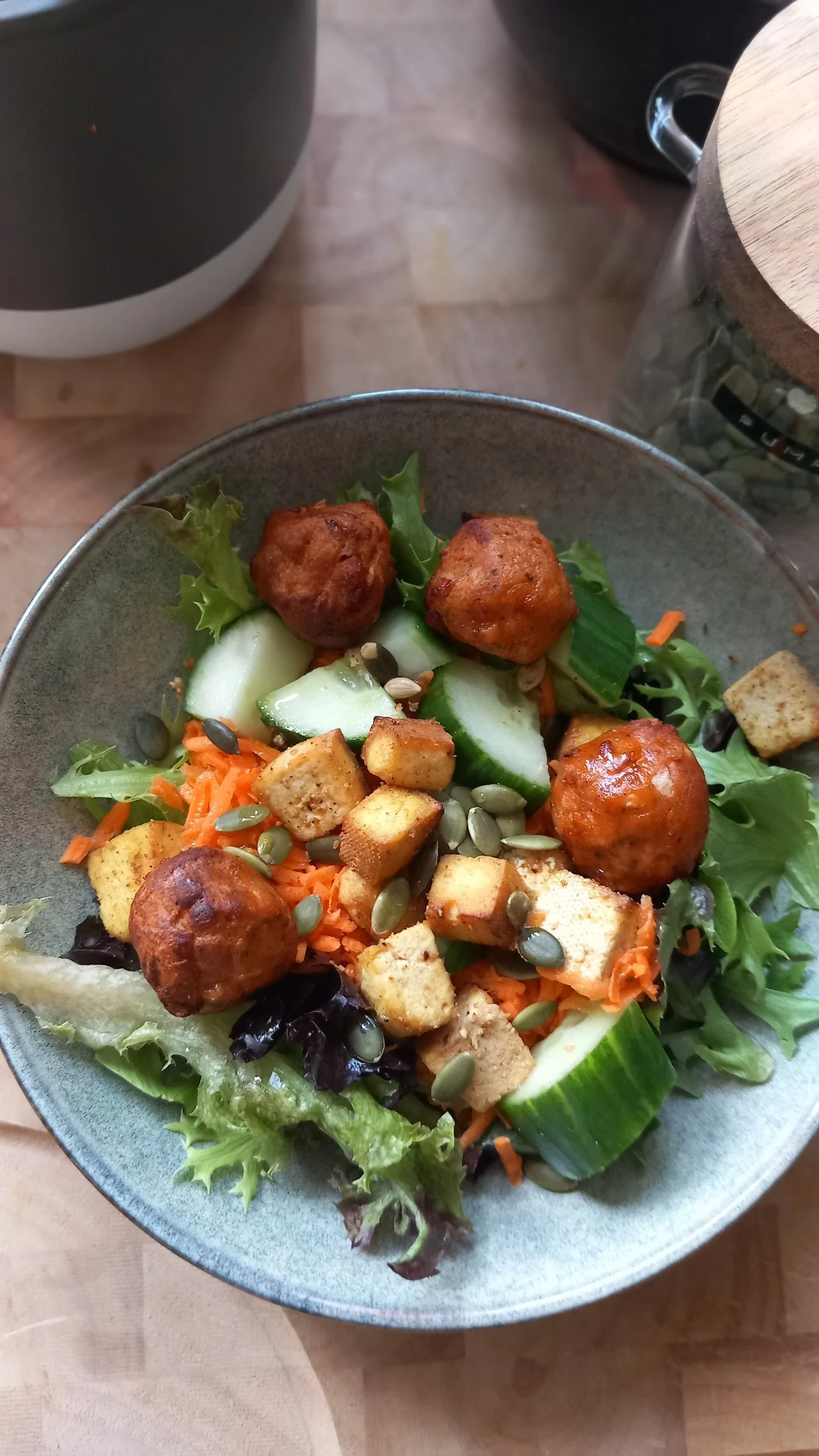 A bowl of salad with lettuce, cucumber, carrots, croutons, pumpkin seeds, and meatballs.