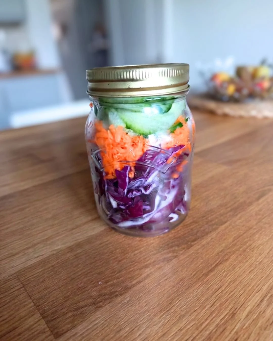 A glass jar filled with sliced cucumbers, shredded carrots, and purple cabbage on a wooden table.