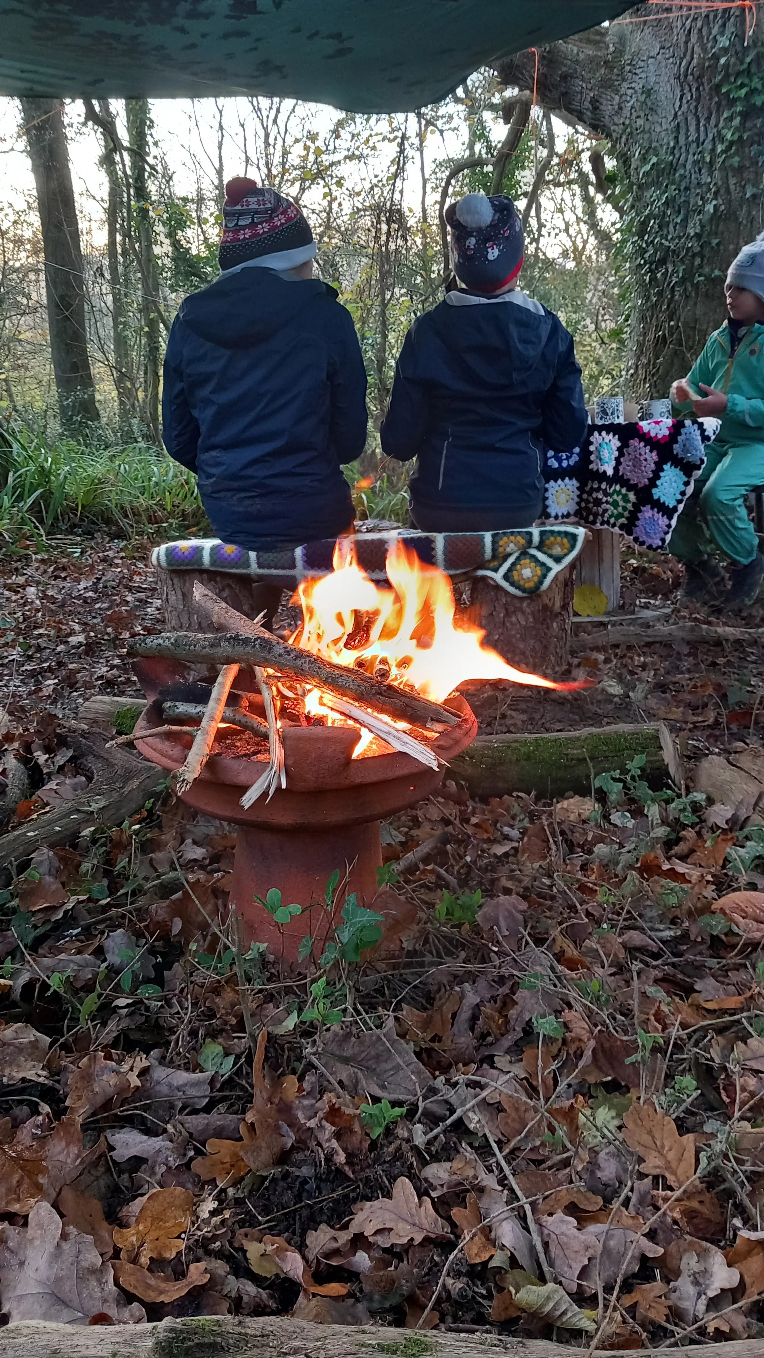 Three children are sitting on logs around a campfire in a wooded area during the evening, with some blankets and a table with crafts nearby.
