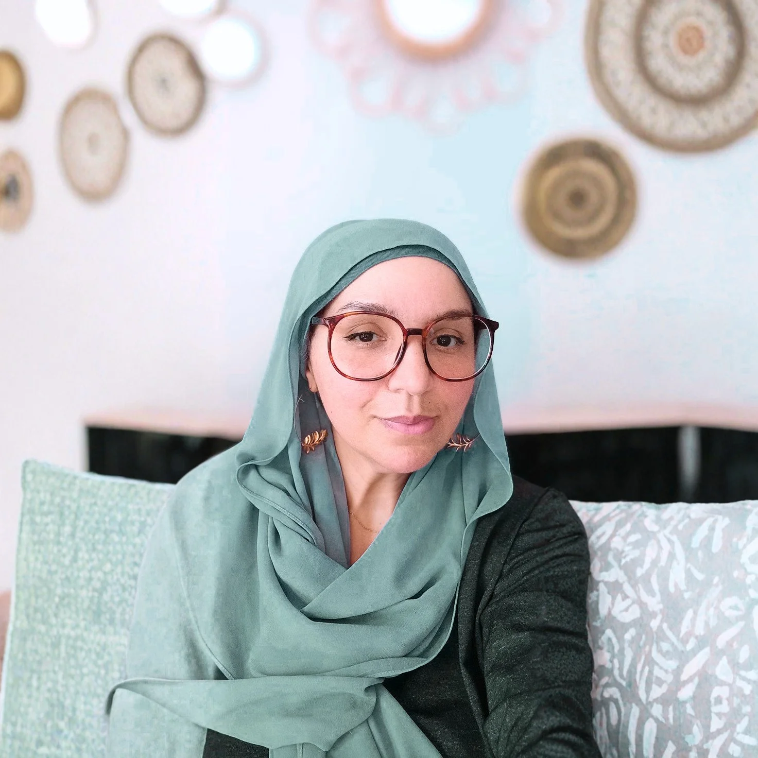 A woman wearing glasses and a green hijab, sitting on a patterned cushion with a light-colored pillow behind her, with decorative circular wall art in the background.