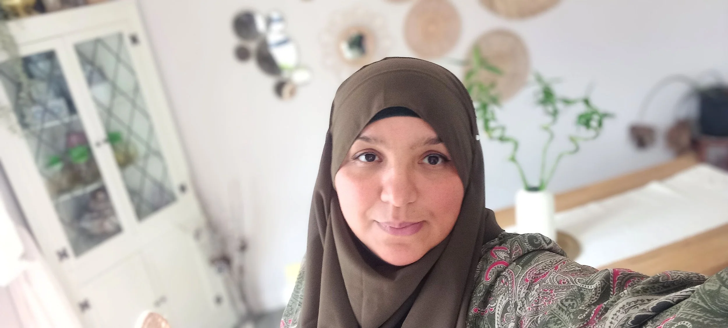 A woman wearing a brown hijab taking a selfie in a home interior, with a white cabinet, a plant, and decorative wall hangings in the background.