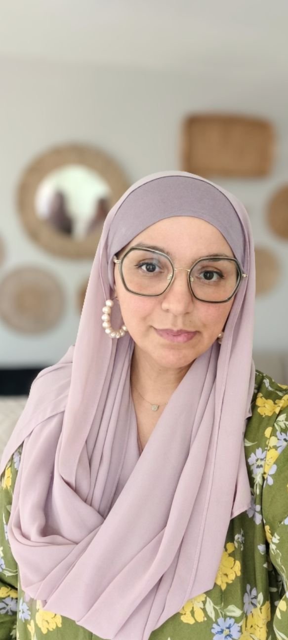 A woman wearing a lavender hijab, glasses, and earrings with pearls, taking a selfie in a room with circle mirrors on the wall.