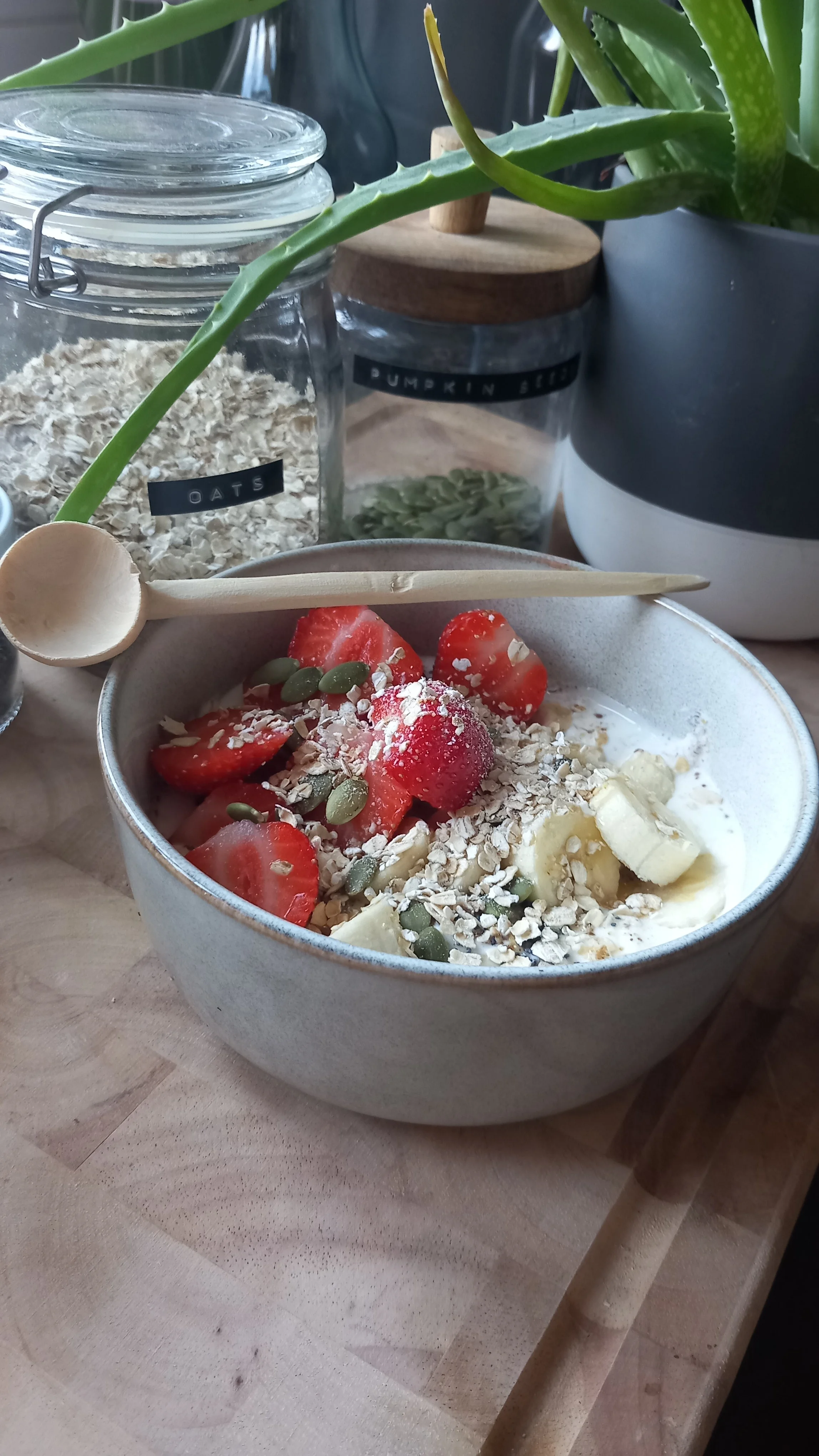 A bowl of yogurt topped with sliced strawberries, banana slices, chopped nuts, and pumpkin seeds on a wooden table. In the background, there are glass jars labeled oats and pumpkin seeds, and a potted aloe vera plant.