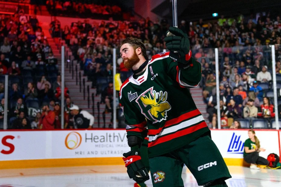 A hockey player in a green jersey with a moose logo on the front stands on the ice rink during a game, with a large crowd of spectators in the background.
