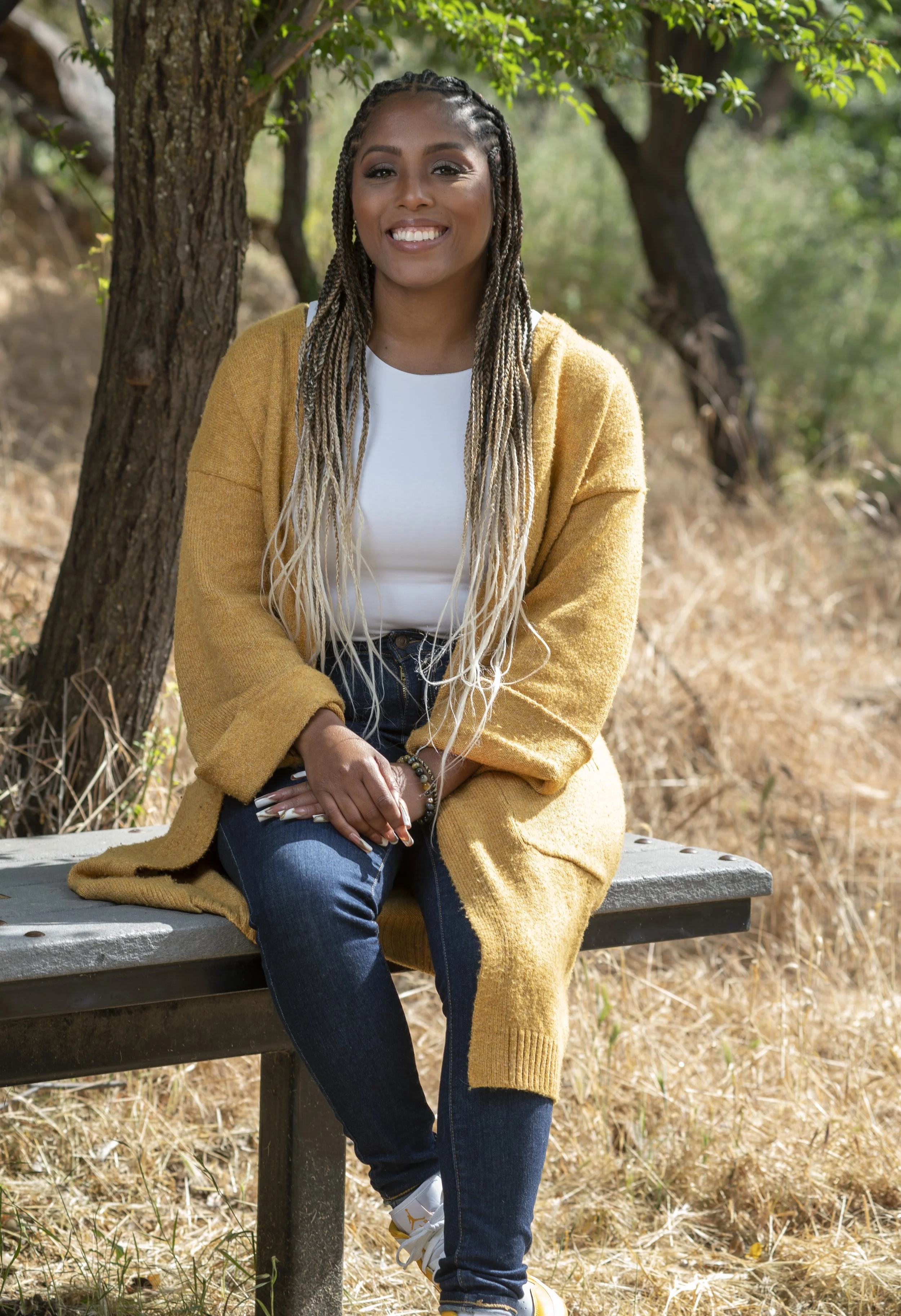 A smiling woman with long braids, wearing a yellow cardigan, white top, and jeans, sitting on a park bench in a natural outdoor setting with trees and dry grass.