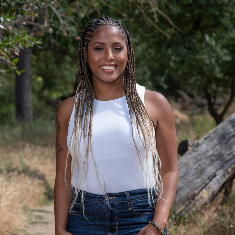 A woman with braided hair, wearing a white sleeveless top and jeans, smiling outdoors in a natural setting with trees and a fallen log in the background.