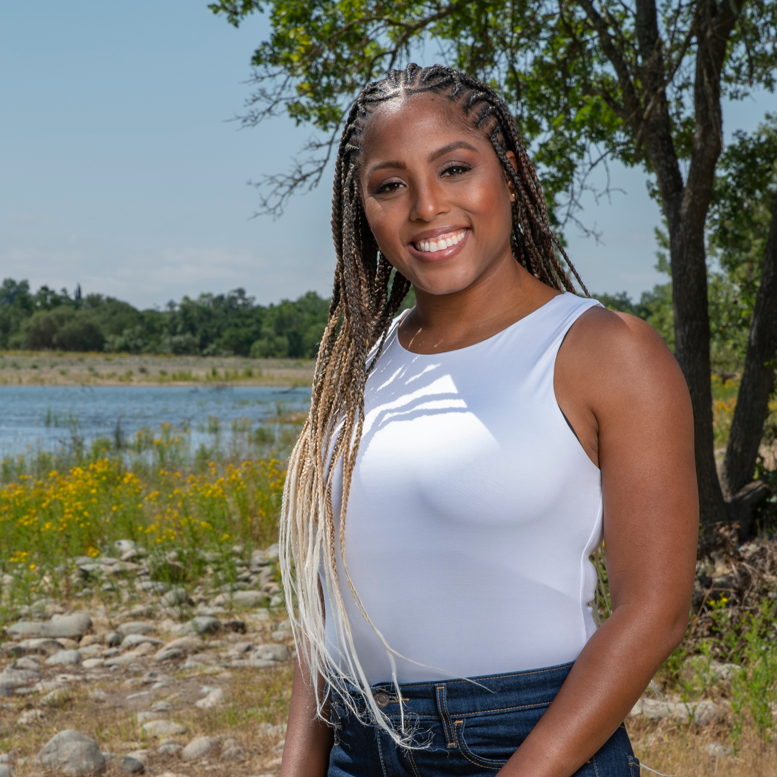 A  BIPOC/ Black young woman with braids smiling outdoors near a body of water, with trees and yellow flowers in the background.