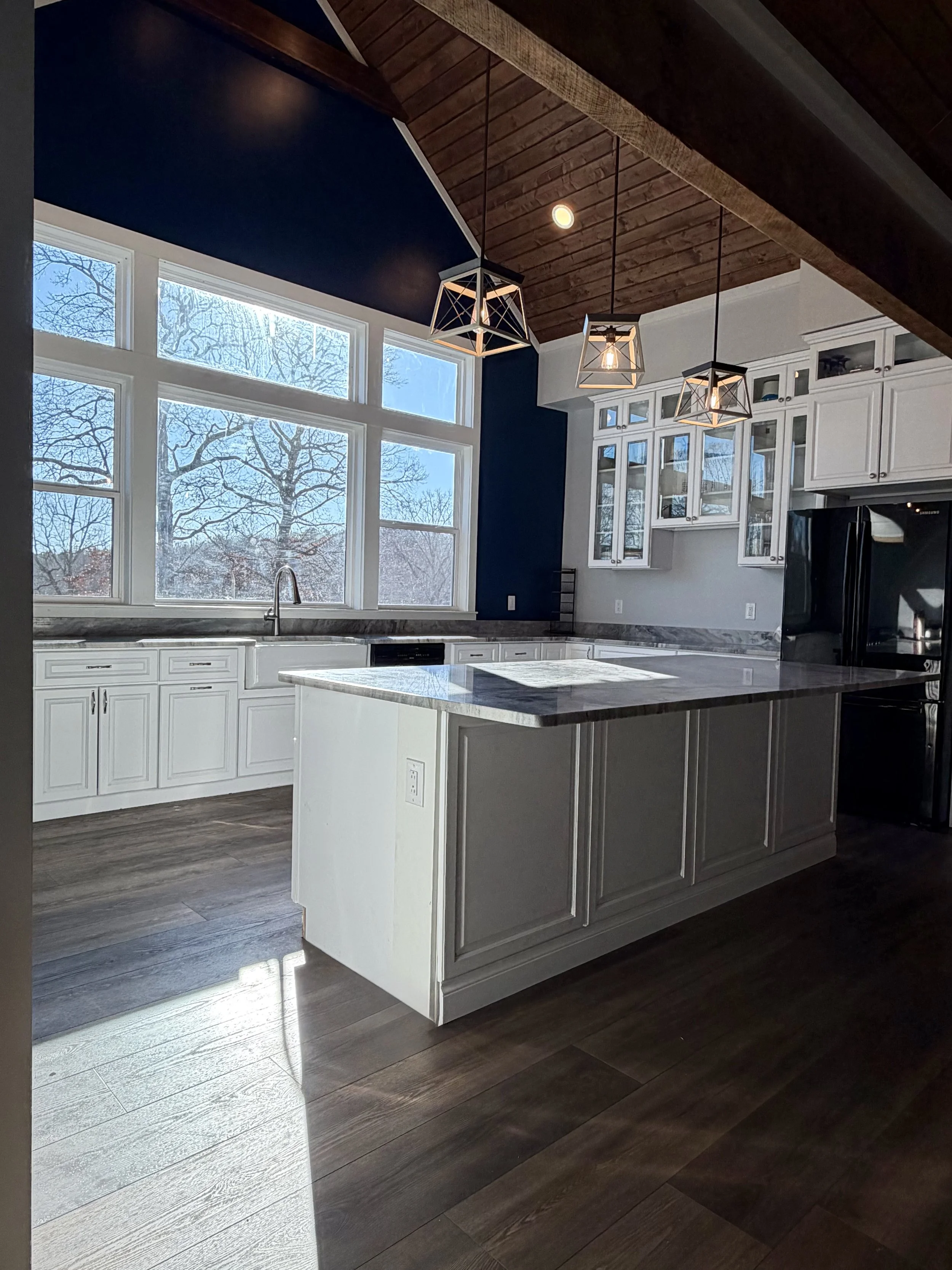 Modern kitchen with large windows, white cabinets, a kitchen island with a marble countertop, dark hardwood flooring, and geometric pendant lights hanging from a wooden ceiling.