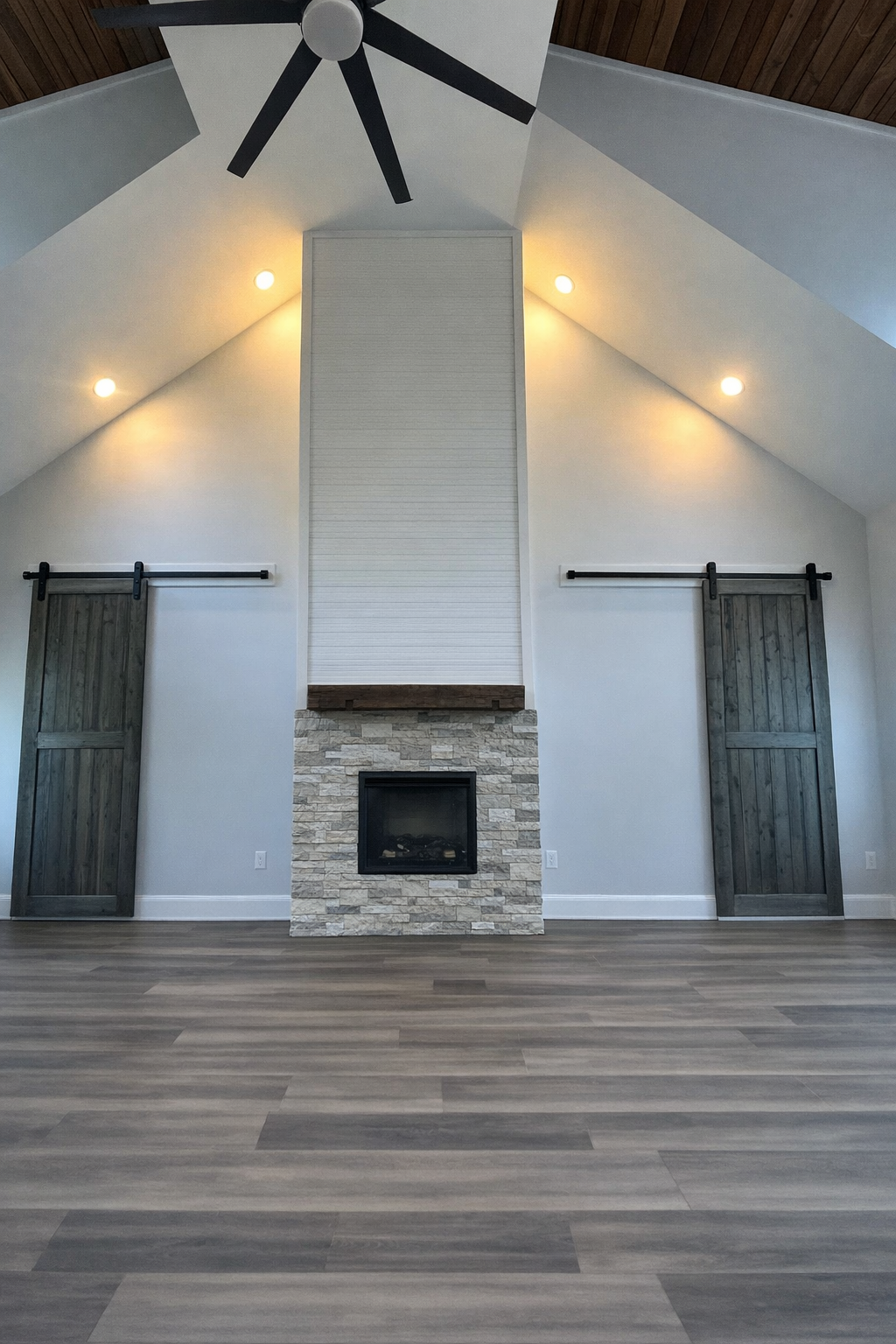 Interior view of a modern living room with a stone fireplace, white walls, wood sliding barn doors, and a vaulted ceiling with a ceiling fan.