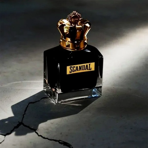 A black perfume bottle labeled 'SCANDAL' with a gold crown-shaped cap, casting a shadow with a crack on the surface it's placed on.