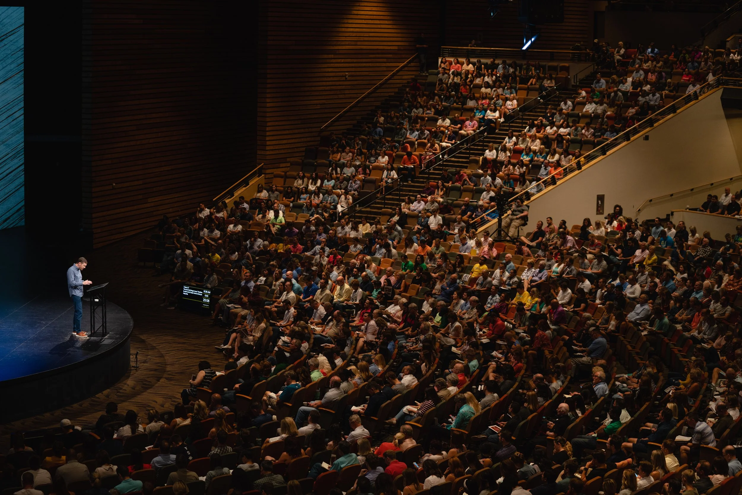 A large auditorium filled with a diverse audience watching a speaker at a podium on stage.