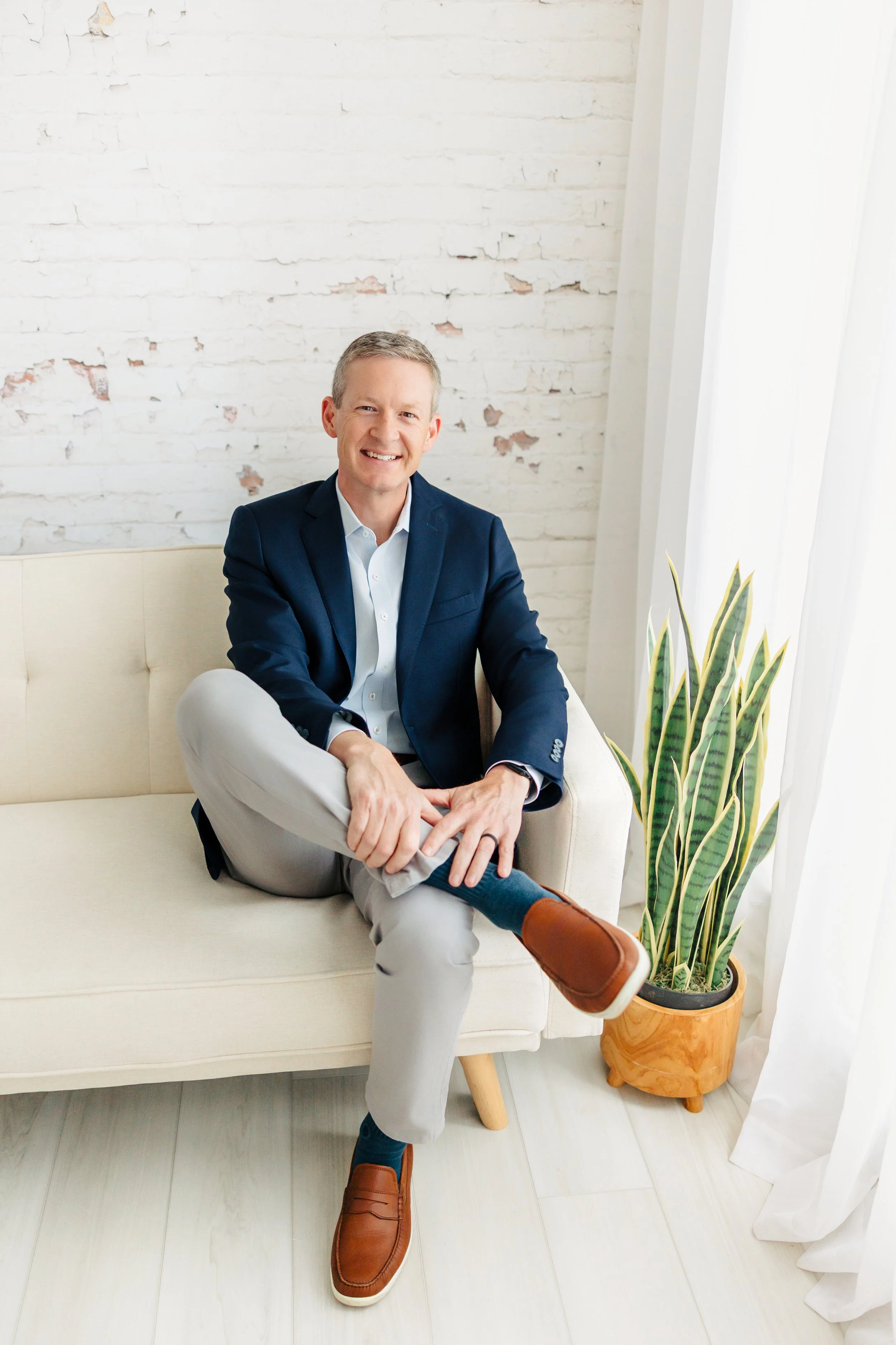Smiling man in blue blazer and beige pants sitting on a cream-colored sofa next to a potted snake plant, with a white brick wall and sheer curtains behind him.